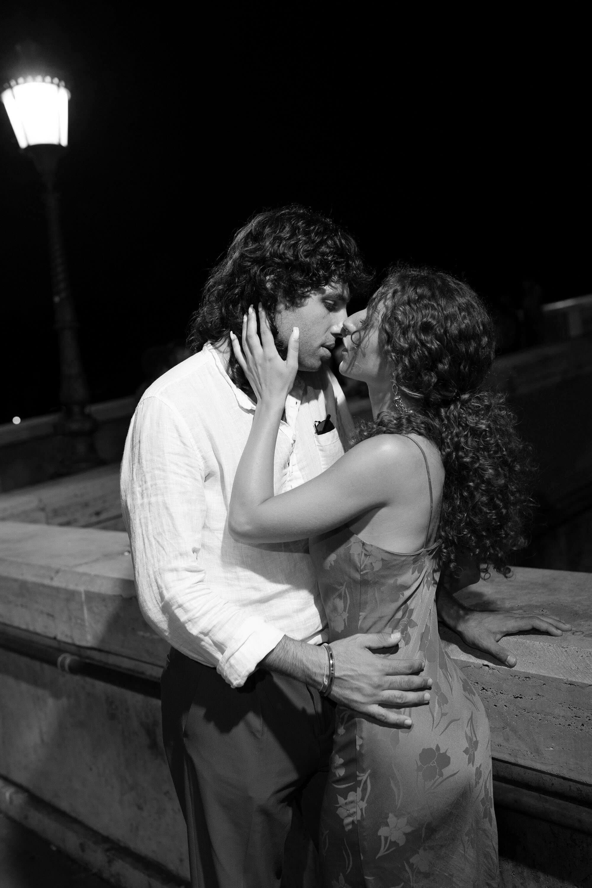 Cinematic black-and-white photo of a couple kissing under a street lamp at night by Sabra Photo
