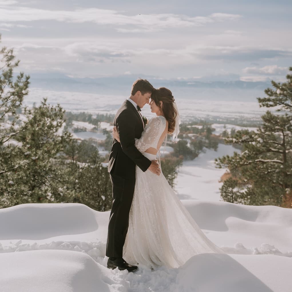 Couple embracing in fresh snow with a sweeping mountain panorama behind them at Sanctuary Golf Course in Sedalia, Colorado